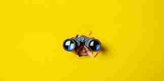 Spyware concept hand holds black binoculars on a yellow background.
